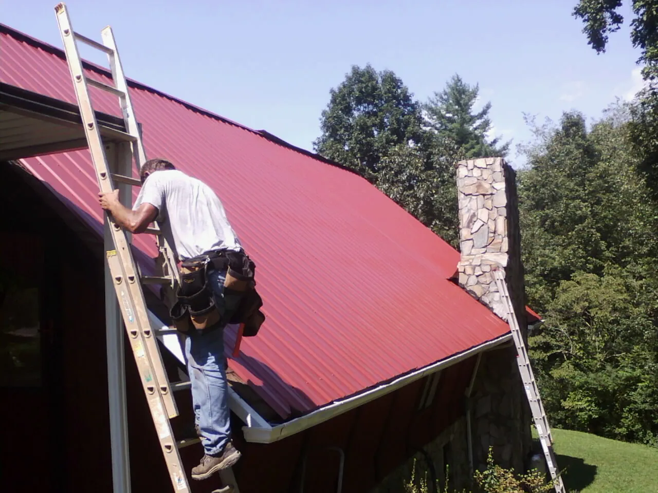 Roofer on a ladder beside a red metal roof and stone chimney — Soffit & Fascia Repair by Roofing Brothers in Aubrey